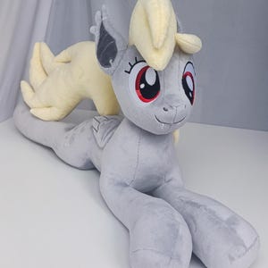Puede incluir: Un peluche de pony en posición tumbada, con cuerpo gris, melena y cola amarillo claro, y gran cara con ojos rojos. El juguete tiene orejas de murciélago y una expresión amigable, ideal para un dormitorio infantil.