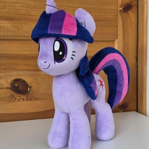 May include: A plush toy of a purple pony character with a horn, large dark eyes, and a mane featuring dark blue and pink stripes. The pony has a dark blue and pink striped tail and a small red star emblem on its flank, standing upright.