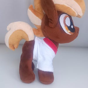 May include: A brown and tan plush toy horse with a white shirt and a red collar. The horse has a brown body, tan mane and tail, and a white shirt with a red collar.