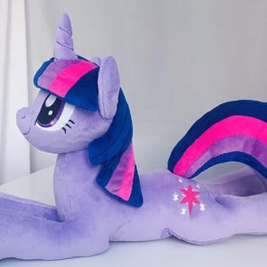 Könnte beinhalten: Ein flauschiges, lavendelfarbenes Einhorn-Spielzeug mit blauer und rosa Mähne und Schweif. Das Einhorn hat ein weißes Sternen-Emblem an der Seite und ein Horn. Das Spielzeug liegt auf einer weißen Oberfläche.