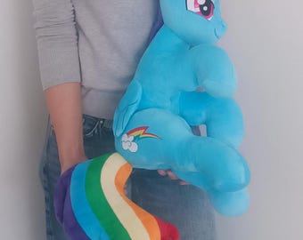 May include: A large, plush, blue pony toy with a rainbow tail and a red, orange, and yellow mane. The toy has a smiling face with pink eyes and a rainbow graphic on its side. The toy is being held by a person.
