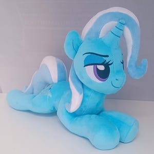 May include: A plush, light blue unicorn toy with a white and blue mane and tail. The toy has purple eyes and is lying down on a white surface. The unicorn is a character from the My Little Pony franchise.