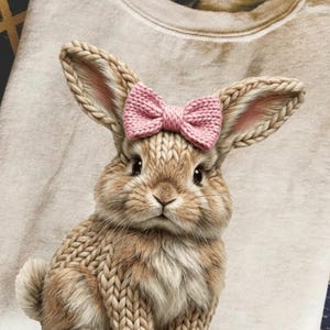 May include: A beige sweatshirt featuring a detailed illustration of a bunny with a pink bow. The bunny has knitted-style fur and long ears with pink inner ears. The sweatshirt is folded on a patterned surface.
