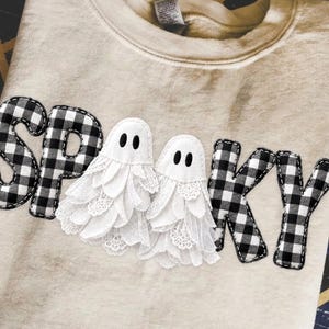 May include: A beige crewneck sweatshirt featuring the word SPOOKY appliqued across the front. The letters S, P, K, and Y are made of black and white buffalo plaid fabric, while two white ghost figures with lace details replace the letter O.