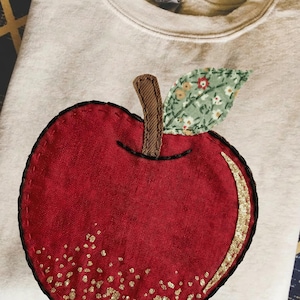 May include: Cream-coloured sweatshirt with a large, appliquéd red apple outlined in black. The apple has a gold glitter accent and a green floral leaf. The stem is brown. The sweatshirt is folded on a patterned surface.