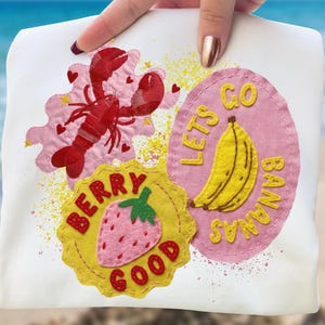 Puede incluir: Tela blanca con tres parches coloridos. Una langosta roja sobre una nube rosa con corazones, un plátano amarillo con el texto "Lets Go Bananas" y una fresa con el texto "Berry Good" en un círculo amarillo.