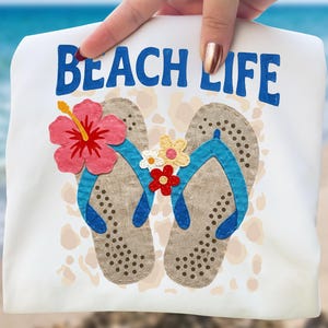 Puede incluir: Toalla de playa blanca con las palabras "BEACH LIFE" en azul. La toalla presenta un diseño de dos chanclas con correas azules, decoradas con flores rosas, amarillas y rojas. El fondo es una escena de playa borrosa.