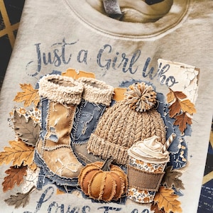 Pode incluir: Camisola bege com um design gráfico com elementos de outono. O design inclui botas, um gorro de malha, uma abóbora e uma chávena de café. O texto "Just a Girl Who Loves Fall" está impresso acima.