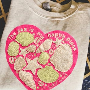 May include: A cream-colored sweatshirt featuring a large pink heart design. The heart is adorned with embroidered seashells, starfish, and the phrase "the sea is my happy place." The text "kukie beach" is also included.