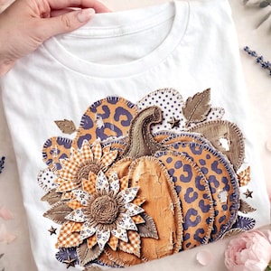 以下が含まれることがあります： 秋をテーマにしたデザインの白いTシャツ。オレンジと紫のヒョウ柄のアクセント、ヒマワリ、葉っぱが特徴です。柔らかい素材で作られたTシャツは、秋に最適です。