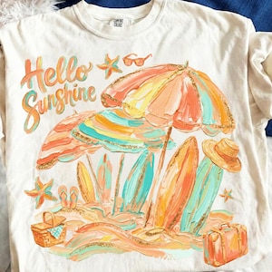 May include: Cream-colored t-shirt with a beach-themed graphic. The design features the words "Hello Sunshine" in orange script, along with beach umbrellas, surfboards, and a suitcase. The artwork uses shades of orange, teal, and yellow.