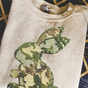 May include: A beige sweatshirt featuring a sequined camouflage bunny design. The bunny is in shades of green, brown, and gold. The sweatshirt is folded neatly, showcasing the bunny's details.