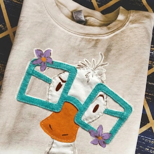 May include: Cream-colored sweatshirt featuring an appliqued duck wearing teal square glasses and purple flowers. The duck has an orange beak and brown eyes. The sweatshirt is folded and appears to be made of a soft material.