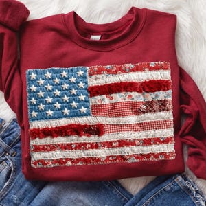Op de afbeelding: Een bordeauxrode sweatshirt met een patchwork Amerikaans vlagontwerp. De vlag heeft geborduurde witte sterren op een blauw veld en rood-witte strepen van verschillende stoffen. De sweatshirt wordt getoond op een wit, bontachtig oppervlak met een blauwe jeans.