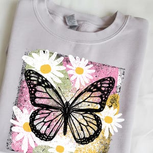 Peut inclure: Un sweat-shirt gris clair avec un motif de papillon et de marguerites. Le papillon pr&eacute;sente des d&eacute;tails noirs et roses, sur un fond rose, vert et jaune. Des marguerites blanches avec des centres jaunes entourent le papillon.