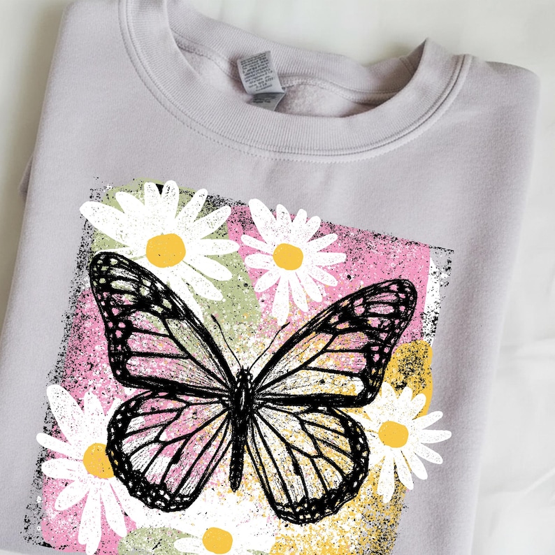 Peut inclure: Un sweat-shirt gris clair avec un motif de papillon et de marguerites. Le papillon pr&eacute;sente des d&eacute;tails noirs et roses, sur un fond rose, vert et jaune. Des marguerites blanches avec des centres jaunes entourent le papillon.