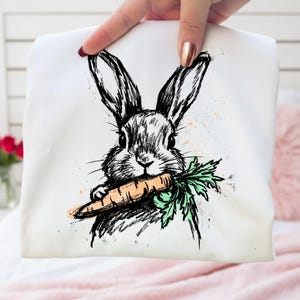 May include: White tote bag featuring a black and white sketch of a rabbit holding a carrot. The rabbit has long ears and is drawn in a detailed, artistic style. The carrot is orange with green leaves, and there are orange paint splatters.