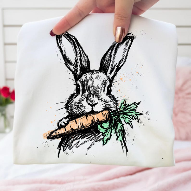 May include: White tote bag featuring a black and white sketch of a rabbit holding a carrot. The rabbit has long ears and is drawn in a detailed, artistic style. The carrot is orange with green leaves, and there are orange paint splatters.