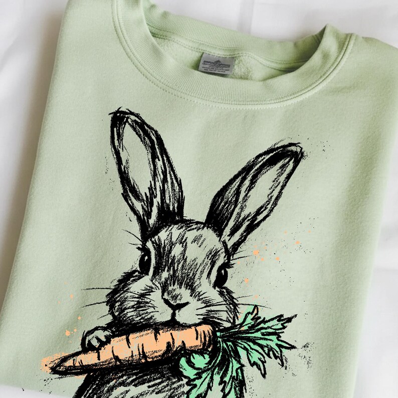May include: A light green sweatshirt featuring a black and white sketch of a rabbit holding a carrot. The carrot is orange with green leaves. The sweatshirt has a crew neck and long sleeves.
