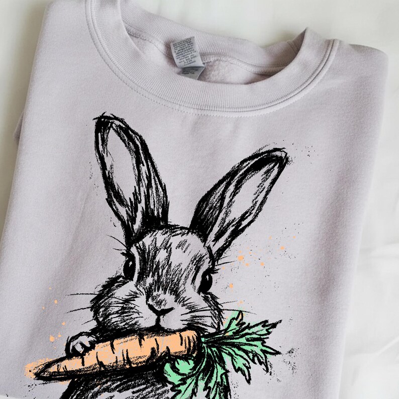 May include: A light purple sweatshirt featuring a black and white sketch of a rabbit holding a carrot. The carrot is orange with green leaves. The design has a hand-drawn aesthetic with paint splatters.