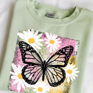 Peut inclure: Un sweat-shirt vert clair avec un motif de papillon noir et blanc et de marguerites. Le papillon est plac&eacute; sur un fond rectangulaire rose, vert et jaune. Le sweat-shirt est en mati&egrave;re douce.