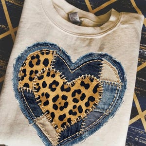 Op de afbeelding: Crèmekleurige sweatshirt met een groot, geplakt hartontwerp. Het hart is samengesteld uit denim, beige stof en luipaardprintstof. De randen van het hart zijn gestikt met een contrasterende draad, wat een unieke, handgemaakte uitstraling creëert.
