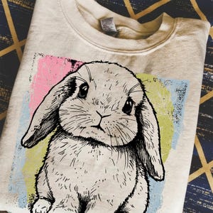 May include: Cream-colored sweatshirt featuring a detailed black and white illustration of a lop-eared bunny. The bunny is set against a colorful backdrop of pink, yellow, and blue. The sweatshirt is folded, showcasing the design prominently.