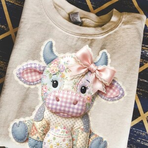 Puede incluir: Una sudadera beige con un diseño de vaca de retazos. La vaca tiene patrones florales y de cuadros, un lazo rosa y detalles de mezclilla azul. El diseño está centrado en la parte delantera de la sudadera.