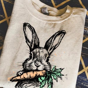 May include: A cream-colored sweatshirt featuring a black and white sketch of a rabbit holding a carrot. The rabbit's face is detailed with expressive eyes and whiskers. The carrot is orange with green leaves.