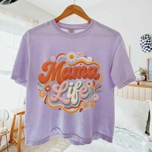 Peut inclure: Un t-shirt lilas avec l'inscription "Mama Life" dans une typographie rétro, entourée de motifs floraux et tourbillonnants colorés. Le t-shirt est suspendu à un cintre en bois.