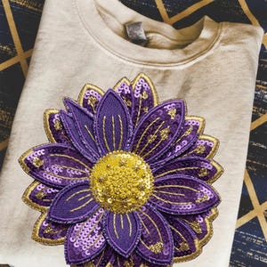 Może przedstawiać: Kremowa bluza z dużym, haftowanym kwiatem. Kwiat ma fioletowe płatki ze złotymi wykończeniami i cekinami oraz żółty, cekinowy środek. Bluza jest złożona na wzorzystej powierzchni.