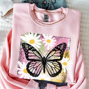 Peut inclure: Un sweat-shirt rose clair orn&eacute; d'un motif de papillon noir et blanc, entour&eacute; de marguerites blanches &agrave; c&oelig;ur jaune. Le motif est encadr&eacute; dans un rectangle aux teintes roses, vertes et jaunes, cr&eacute;ant un contraste saisissant.