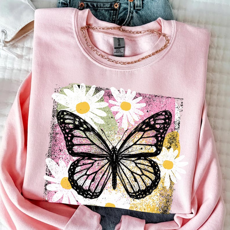 Peut inclure: Un sweat-shirt rose clair orn&eacute; d'un motif de papillon noir et blanc, entour&eacute; de marguerites blanches &agrave; c&oelig;ur jaune. Le motif est encadr&eacute; dans un rectangle aux teintes roses, vertes et jaunes, cr&eacute;ant un contraste saisissant.
