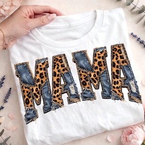 Puede incluir: Camiseta blanca con la palabra "MAMA" en letras grandes y audaces. Las letras presentan un diseño de patchwork que combina mezclilla e impresión de leopardo. La camiseta está doblada y se muestra sobre una superficie clara.
