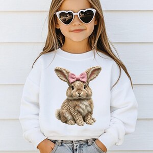 May include: White sweatshirt featuring a detailed illustration of a brown bunny with a pink bow. The bunny has knitted-style fur and long ears. The sweatshirt is a crew neck style.