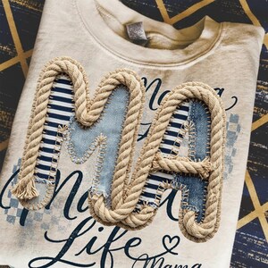 May include: Cream-colored sweatshirt with the letters "MA" appliqued on the front. The letters are made of denim and striped fabric, outlined with rope. The words "No Life Mama" are printed in navy blue script.