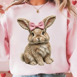 May include: Pink sweatshirt featuring a detailed illustration of a bunny with a pink bow. The bunny has textured fur and is sitting upright. The sweatshirt is a solid light pink color, creating a soft and feminine aesthetic.