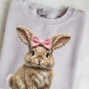 May include: A light purple sweatshirt featuring a detailed illustration of a bunny with knitted-look fur and a pink bow. The bunny has long ears and a fluffy tail, creating a cozy and whimsical design. The sweatshirt is folded neatly.