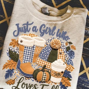 Puede incluir: Una sudadera gris claro con el texto "Just a Girl Who Loves Fall". El diseño incluye un gráfico con temática otoñal con botas, un sombrero, una calabaza y una taza de café. El gráfico está en tonos azules, naranjas y marrones.