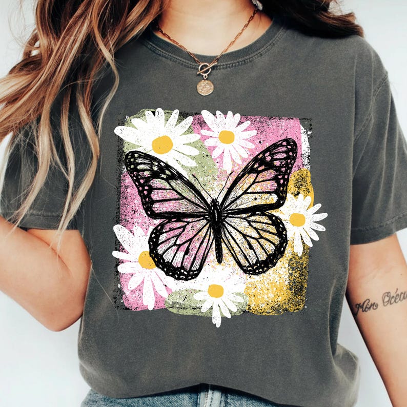Peut inclure: T-shirt gris fonc&eacute; avec un motif de papillon et de marguerites. Le papillon a des ailes noires et blanches avec des accents roses, entour&eacute; de marguerites blanches &agrave; c&oelig;ur jaune. Le motif est sur un fond rose, jaune et vert.