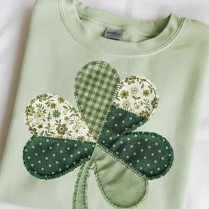 May include: A light green sweatshirt featuring a large, appliqued shamrock. The shamrock is made of various green fabrics, including polka dots, floral patterns, and gingham. The stem is a solid green color, and the design is stitched onto the sweatshirt.