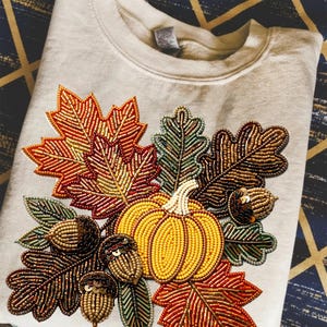Op de afbeelding: Crèmekleurige sweatshirt met een herfstthema kralenontwerp. Het ontwerp toont een gele pompoen, kleurrijke bladeren in tinten rood, oranje en groen en verschillende eikels. De kralen geven textuur en dimensie aan de herfstkleding.