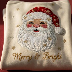 Może przedstawiać: Kremowa bluza z haftem przedstawiającym twarz Świętego Mikołaja, czerwoną czapkę i białą brodę. Napis "Merry & Bright" jest haftowany złotą nicią. Złote płatki śniegu rozrzucone wokół Mikołaja tworzą świąteczny motyw.