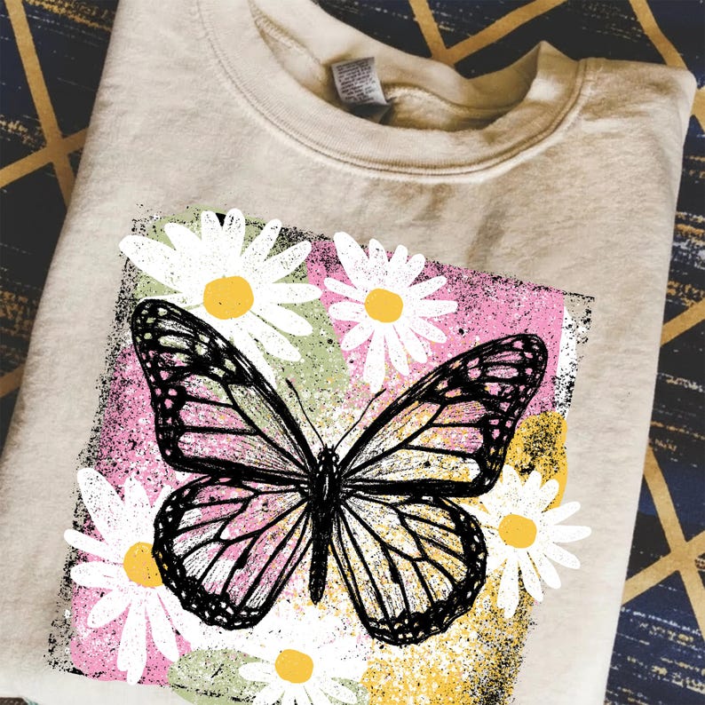 Peut inclure: Un sweat-shirt beige avec un motif de papillon et de marguerites. Le papillon est noir avec des accents roses et jaunes, sur un fond rose, vert et jaune. Des marguerites blanches &agrave; c&oelig;ur jaune entourent le papillon.