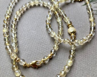 May include: A gold necklace and bracelet set made with clear quartz beads with gold flecks. The necklace has a gold fish charm and the bracelet has a gold ball charm.
