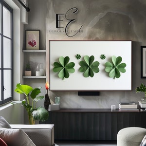 May include: A framed artwork featuring three large green shamrocks and two smaller ones against a white background. The artwork is displayed on a wall with the text "Echoes & Ember" above it. The room has a modern design with plants and a dark cabinet.