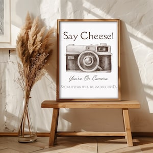 以下が含まれることがあります： ヴィンテージカメラのイラストと「Say Cheese! You're On Camera. Shoplifters Will Be Prosecuted.」というテキストが書かれた額装されたプリント