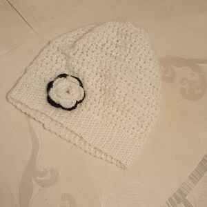 Op de afbeelding: Witte gebreide beanie met een zwart-witte bloemenversiering.