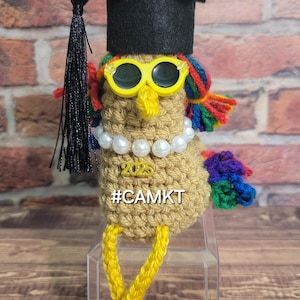 May include: A crocheted bird wearing a black graduation cap, yellow sunglasses, a pearl necklace, and a rainbow tail. The bird is sitting on a clear pedestal and has a yellow "2023" charm on its chest. The text "#CAMKT" is written below the charm.