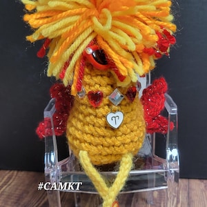 May include: A handmade, crocheted bird figurine with yellow, orange, and red yarn. The bird has a fluffy head, red heart-shaped embellishments, and a small pendant. It is sitting on a clear acrylic chair.